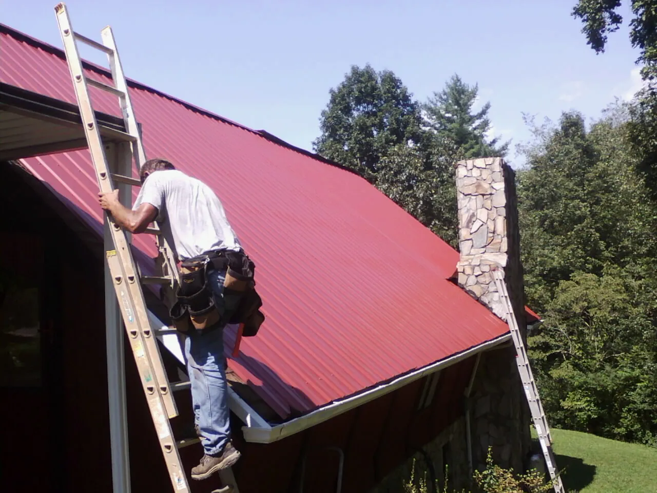 Roofer on a ladder beside a red metal roof and stone chimney — Soffit & Fascia Repair by Roofing Brothers in Pound Ridge